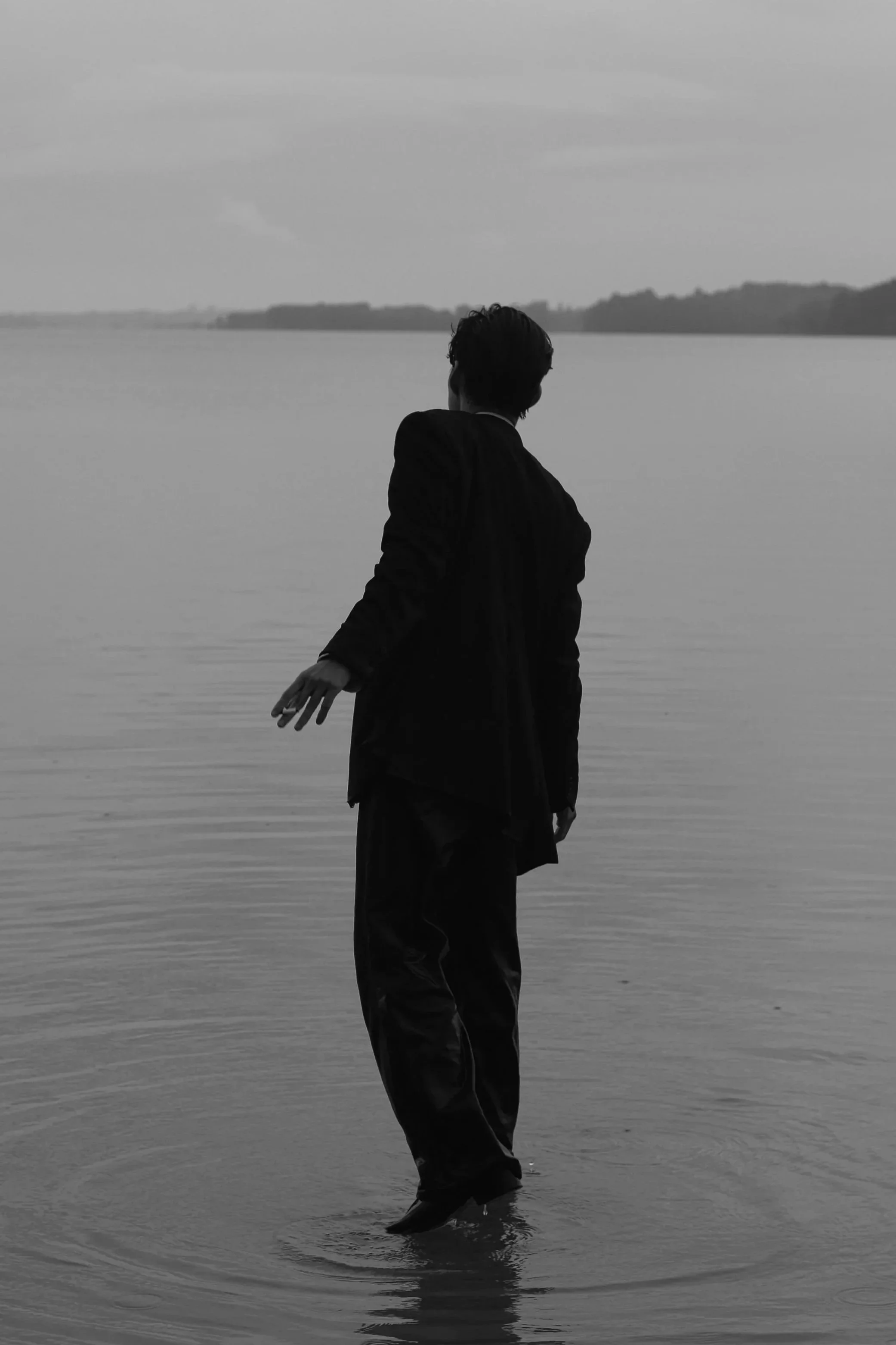 A person in dark clothing standing in shallow water at the edge of a lake, with their back to the camera and face turned away, looking toward distant landmasses under a cloudy sky in black and white.