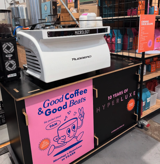 A coffee machine labeled 'Microcology' on a store counter, surrounded by cups and shelves of assorted products, with pink and black promotional signs for Microcology and Hyperluxe.