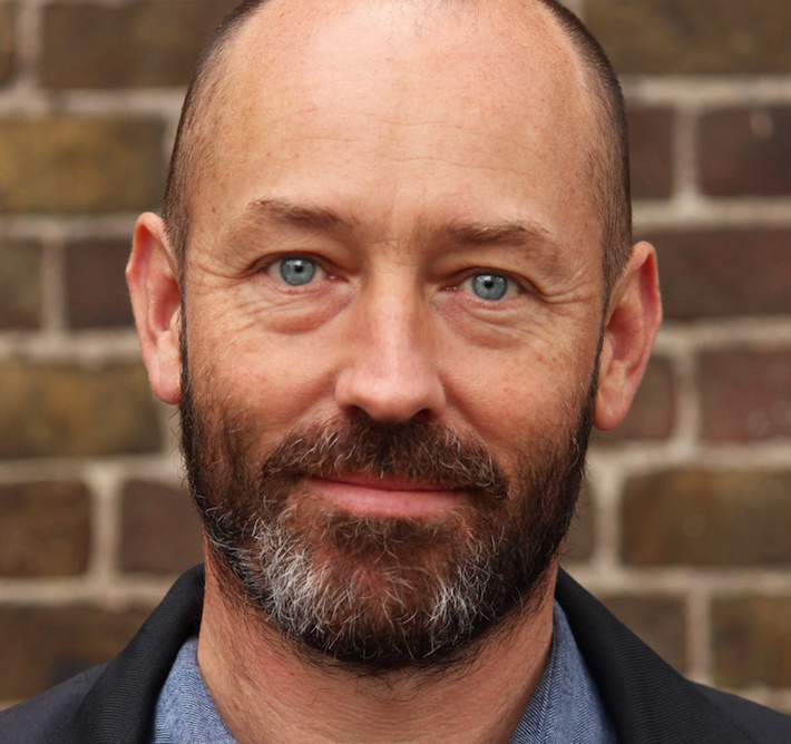 Close-up of a man with a beard and blue eyes, smiling slightly, standing outdoors in front of a brick wall.