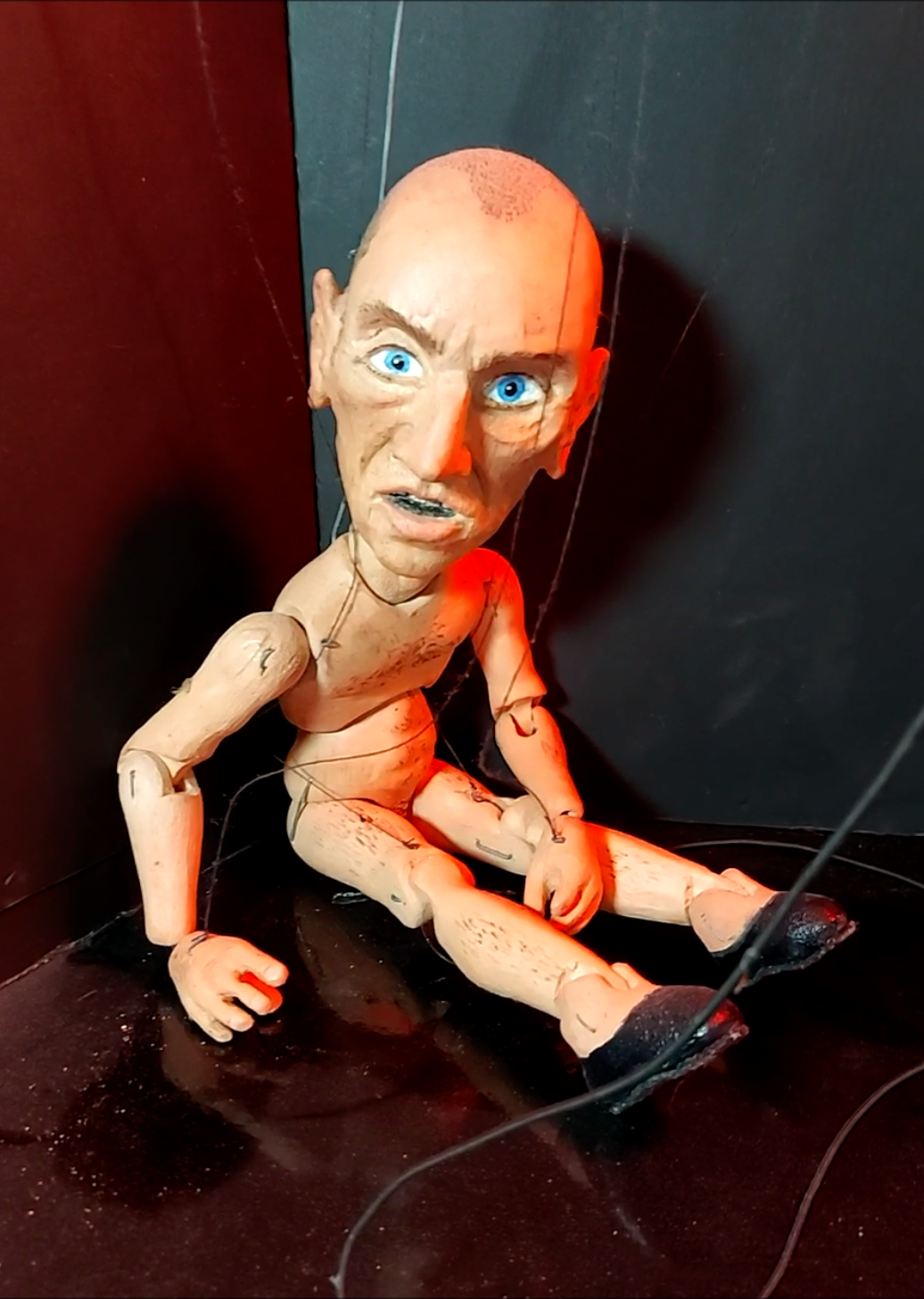 A creepy, worn puppet with a bald head, large blue eyes, and a distressed facial expression, sitting on a dark surface with spider webs and red lighting.