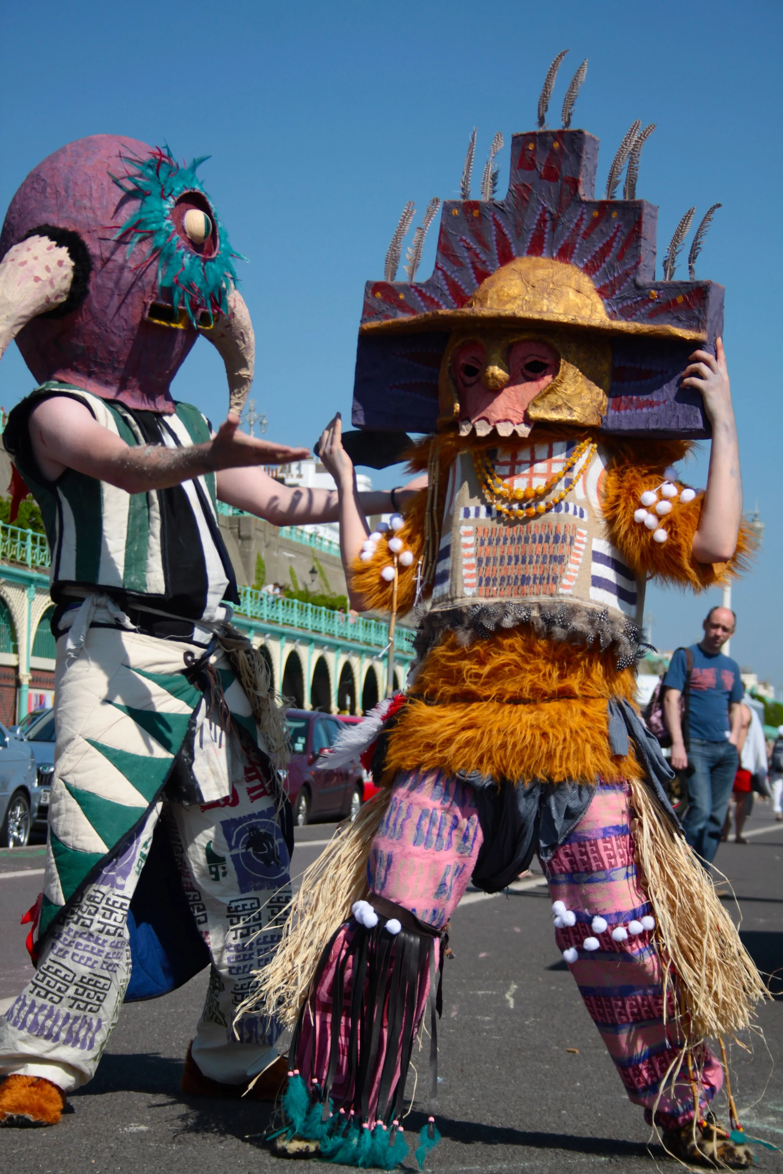 Two people dressed in colorful, elaborate costumes with masks, participating in a parade or festival on a street on a sunny day.