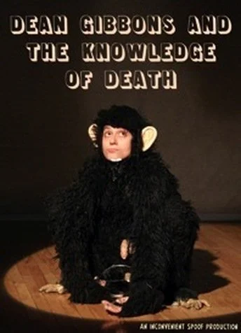 A person dressed as a black monkey costume sitting on a stage, holding a black object. The background has text that reads 'Dean Gibbons and the Knowledge of Death' and 'An Inconvenient Spoon Production.'
