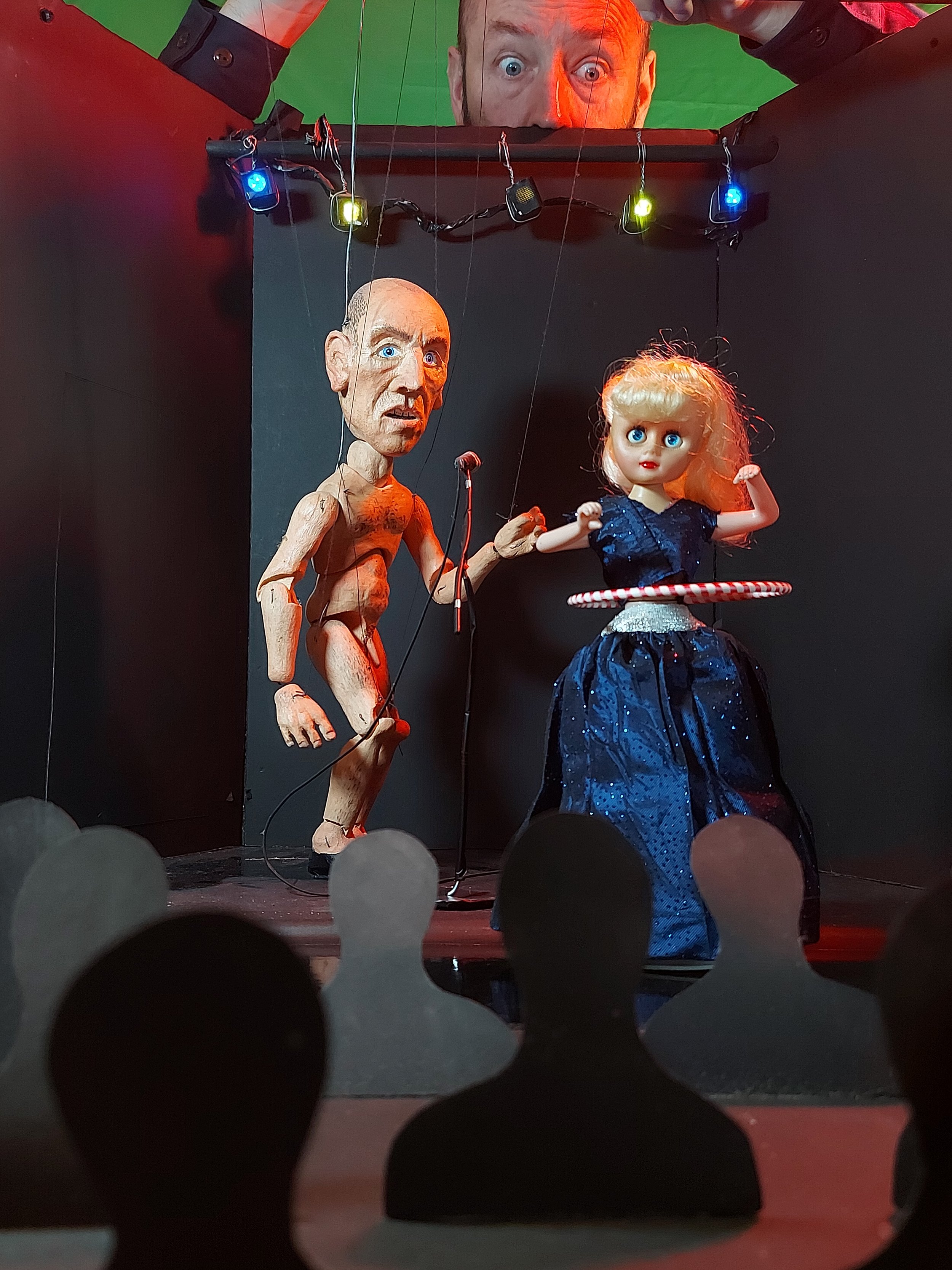 A puppet show features a wooden puppet resembling an old man with a serious expression and blue eyes, and a doll resembling a girl with blonde hair and big blue eyes, dressed in a dark blue sparkly dress with a red-and-white striped hula hoop around 