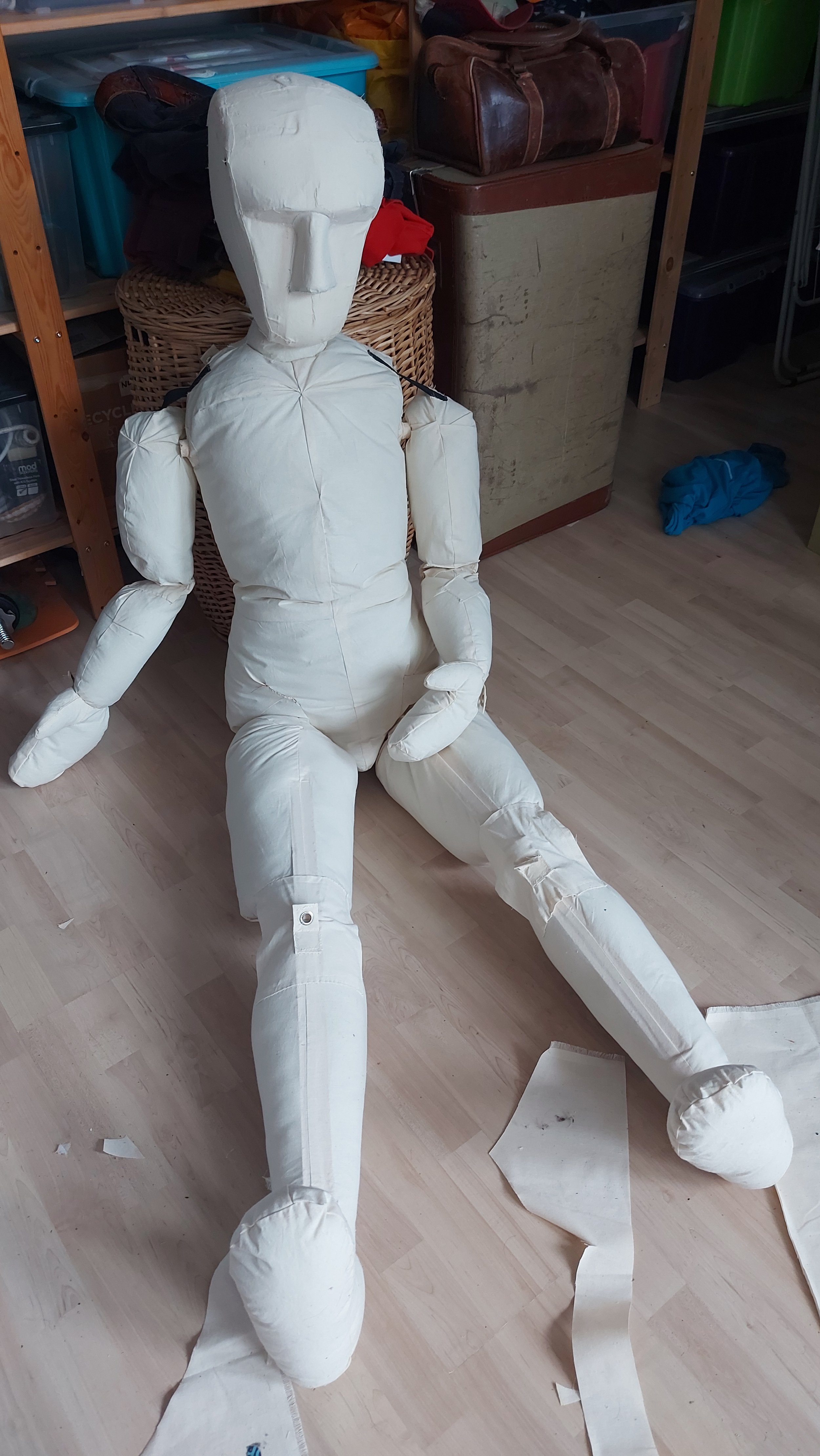 A life-sized puppet or costume resembling a human figure, made of white fabric or padding, seated on the floor in a room with shelves and storage boxes.