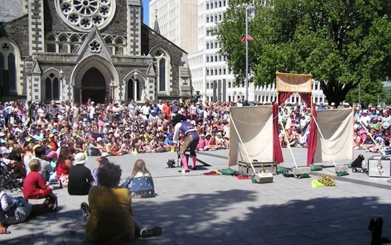 Crowd gathered in front of a street performance set up with handmade puppet theater in front of a historic church in an urban area.