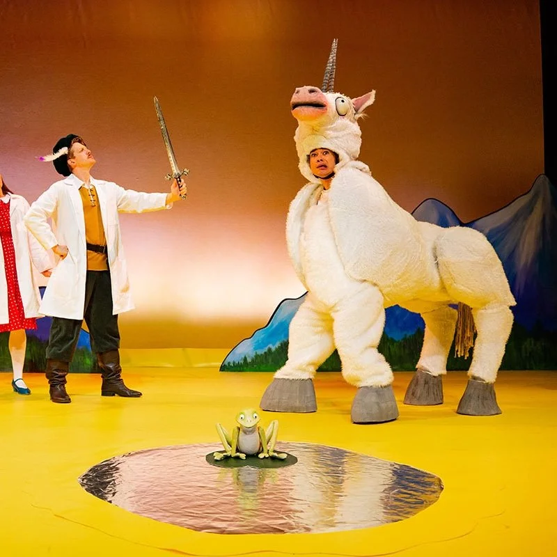 A stage performance featuring people in costumes, including a person in a unicorn costume and another person with a sword, with painted mountains in the background and theatrical scenery.