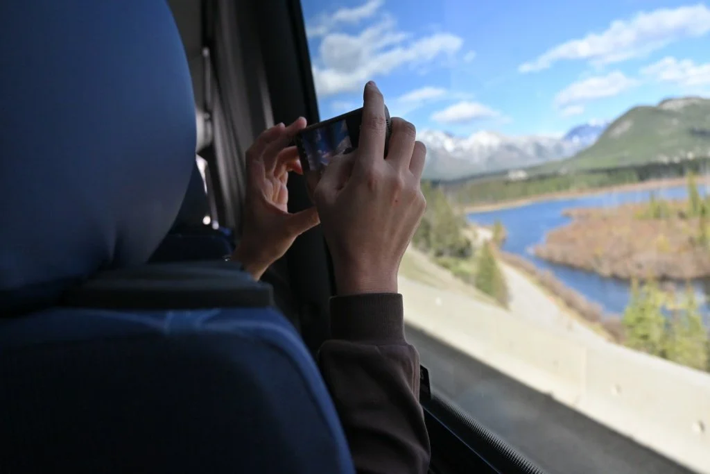Passenger taking a photo with her phone through a bus window, river and mountains visible outside in the Canadian Rockies