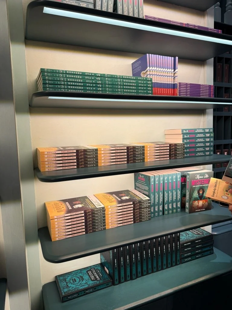 n angled shelf view of Aesop Women's Library 2026 mystery titles, with a hand reaching from the right to pull a yellow-covered book from a lower shelf