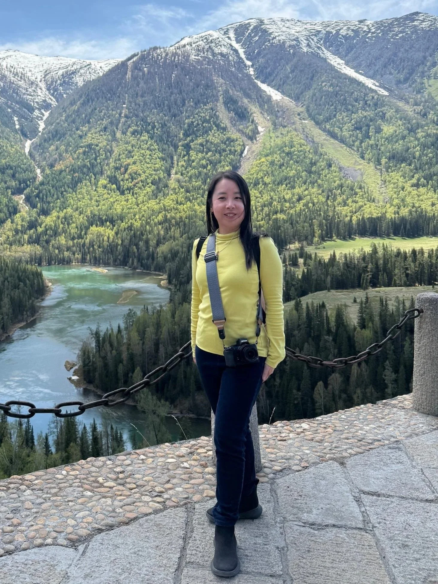 A woman in a yellow long-sleeve shirt and black pants standing on a stone pathway with a scenic mountain and river view in the background.