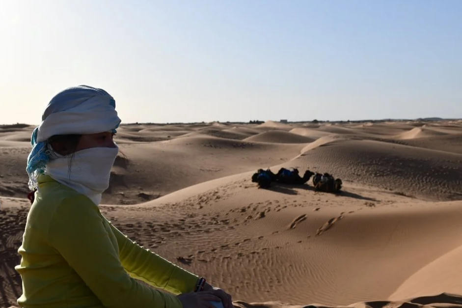 A person wearing a white headscarf and a white face covering sitting in a desert landscape with sand dunes, with a vehicle in the distance.