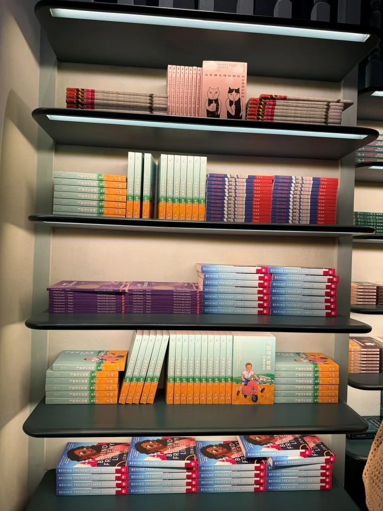 Shelves of Aesop Women's Library 2026 titles including pastel-spined Mandarin children's titles and Behind Frenemy Lines by Zen Cho, with a children's book cover propped face-out