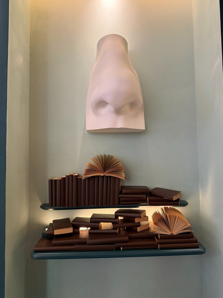 A sculptural plaster nose mounted on a wall above two shelves of decorative faux leather-bound books, one splayed open, at Aesop Women's Library 2026