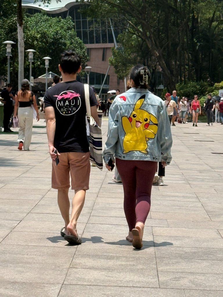 A fan wearing a denim jacket with a hand-painted Pikachu design and Poké Ball patch, walking through KLCC Park during PokéXciting!