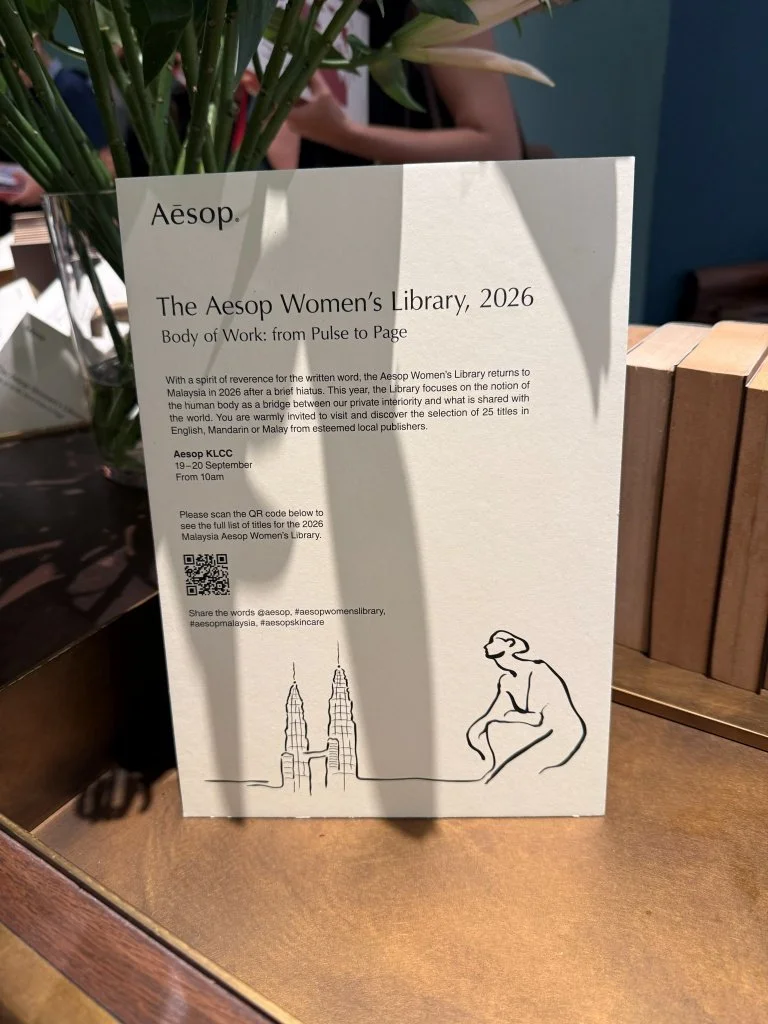 An Aesop Women's Library 2026 information card propped upright beside a vase of lilies, printed with the event theme, dates and a QR code for the full title list.