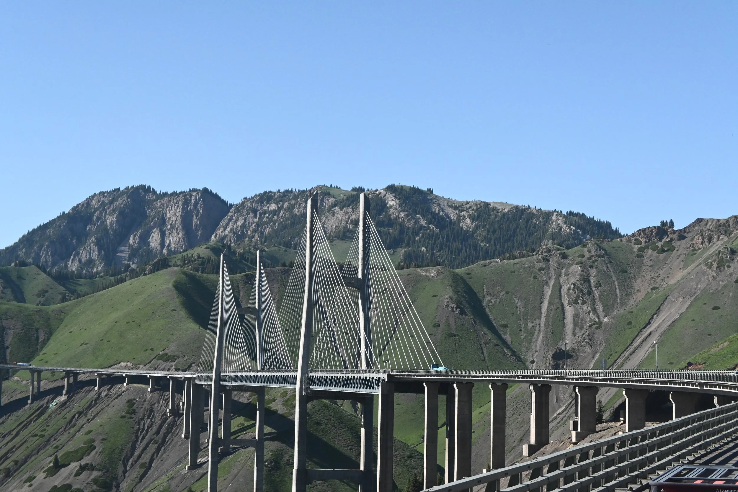 A large cable-stayed bridge with tall pylons and suspension cables spanning across a mountain valley with green slopes and rugged peaks under a clear blue sky.