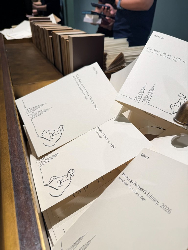 A cluster of Aesop Women's Library 2026 information cards scattered across a wooden counter, with rows of book spines blurred in the background