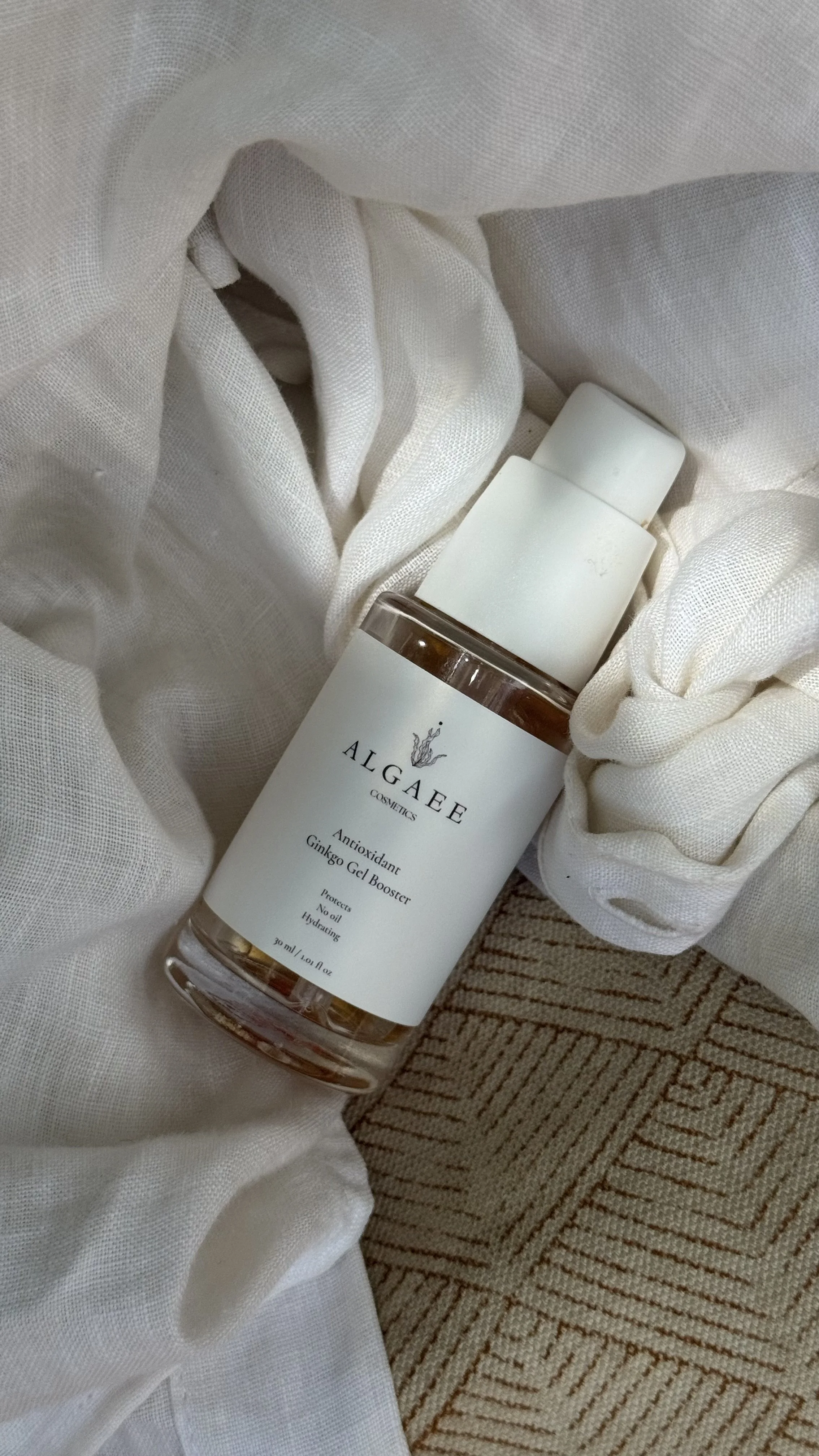 A bottle of ALGEE Cosmetics Antioxidant Ginkgo Gel Booster resting on a beige textured rug, surrounded by white fabric.