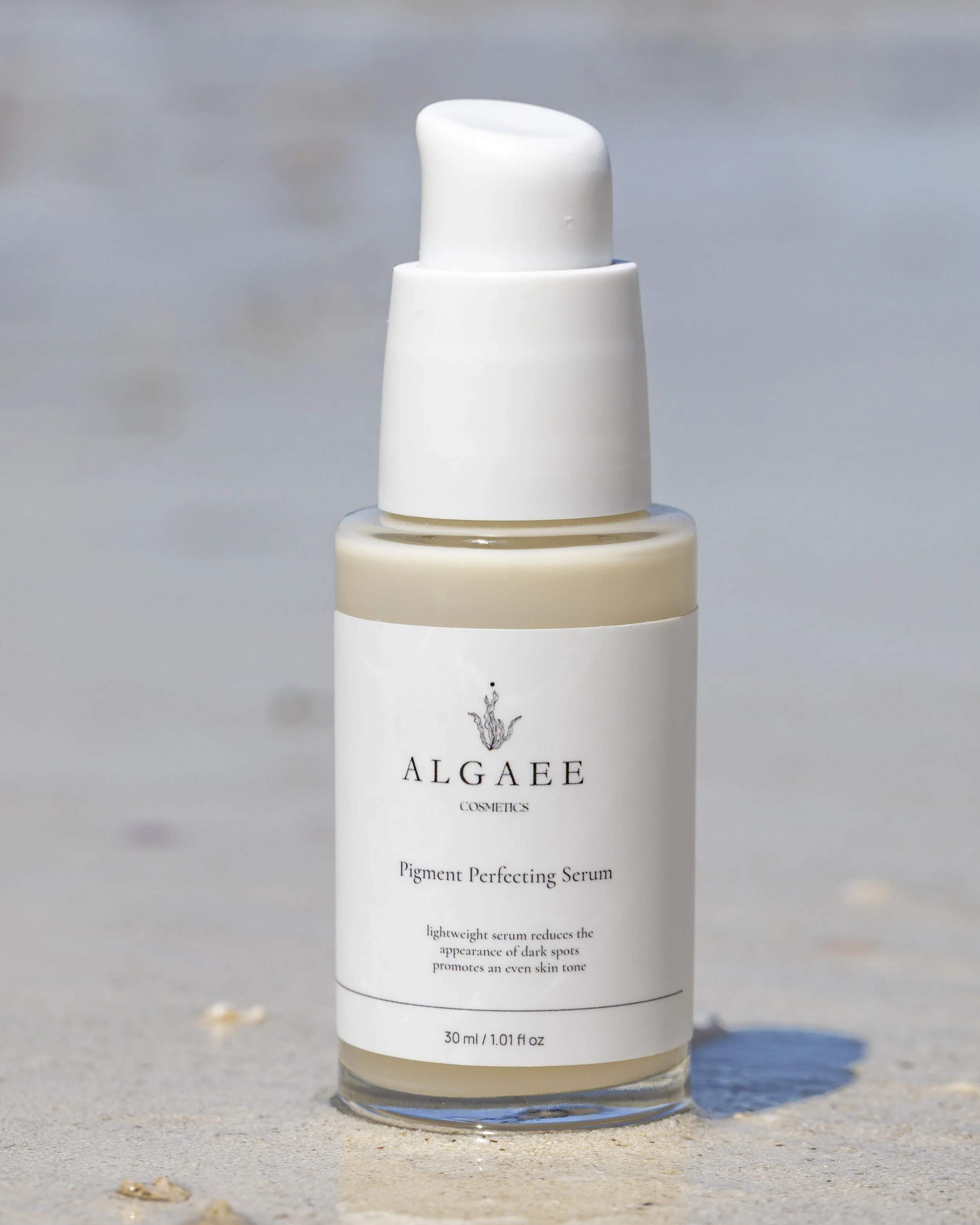 A bottle of Algage Cosmetics Pigment Perfecting Serum standing on a sandy surface near the beach with the ocean and sky in the background.