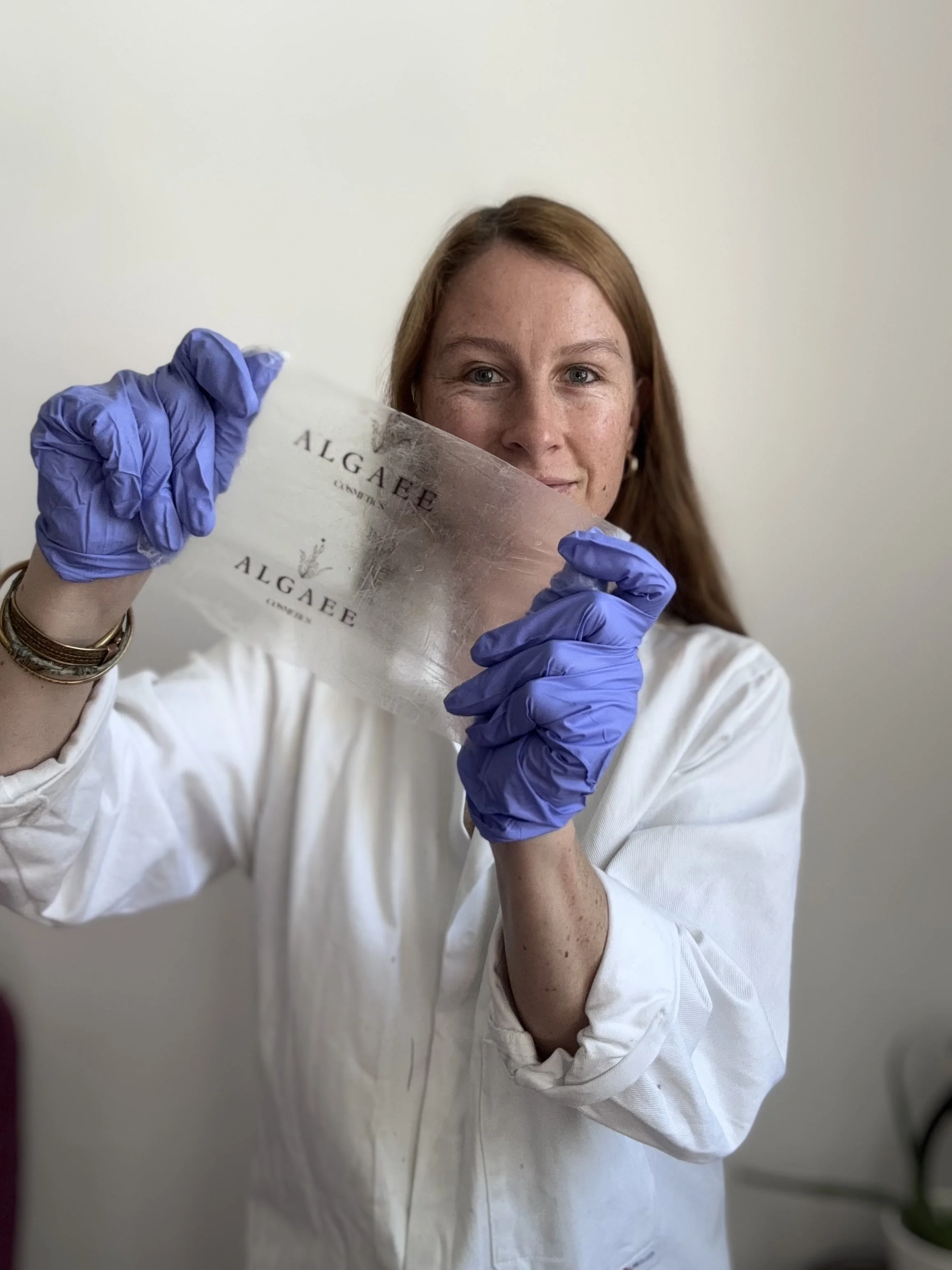 A woman with red hair and light skin is holding a silver-colored plaque that reads 'ALGARBE CALLES' and is wearing purple gloves and a white coat, standing in front of a plain white wall.