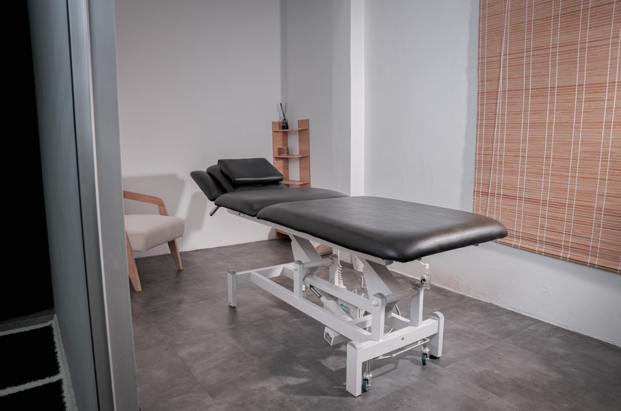 Treatment room with a black massage or examination table in the center, a white chair with wooden arms in the background, and a wooden shelf on the wall with a diffuser or incense holder.