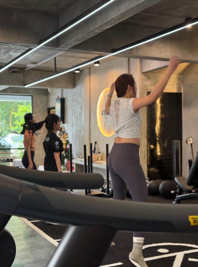 Women working out in a gym with modern, industrial decor. One woman is doing a high kick, others are standing near fitness equipment and a window with greenery outside.