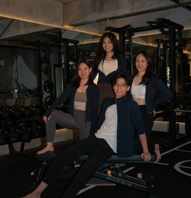 Four young adults, three women and one man, posing in a gym near fitness equipment. They are smiling and dressed in workout attire.