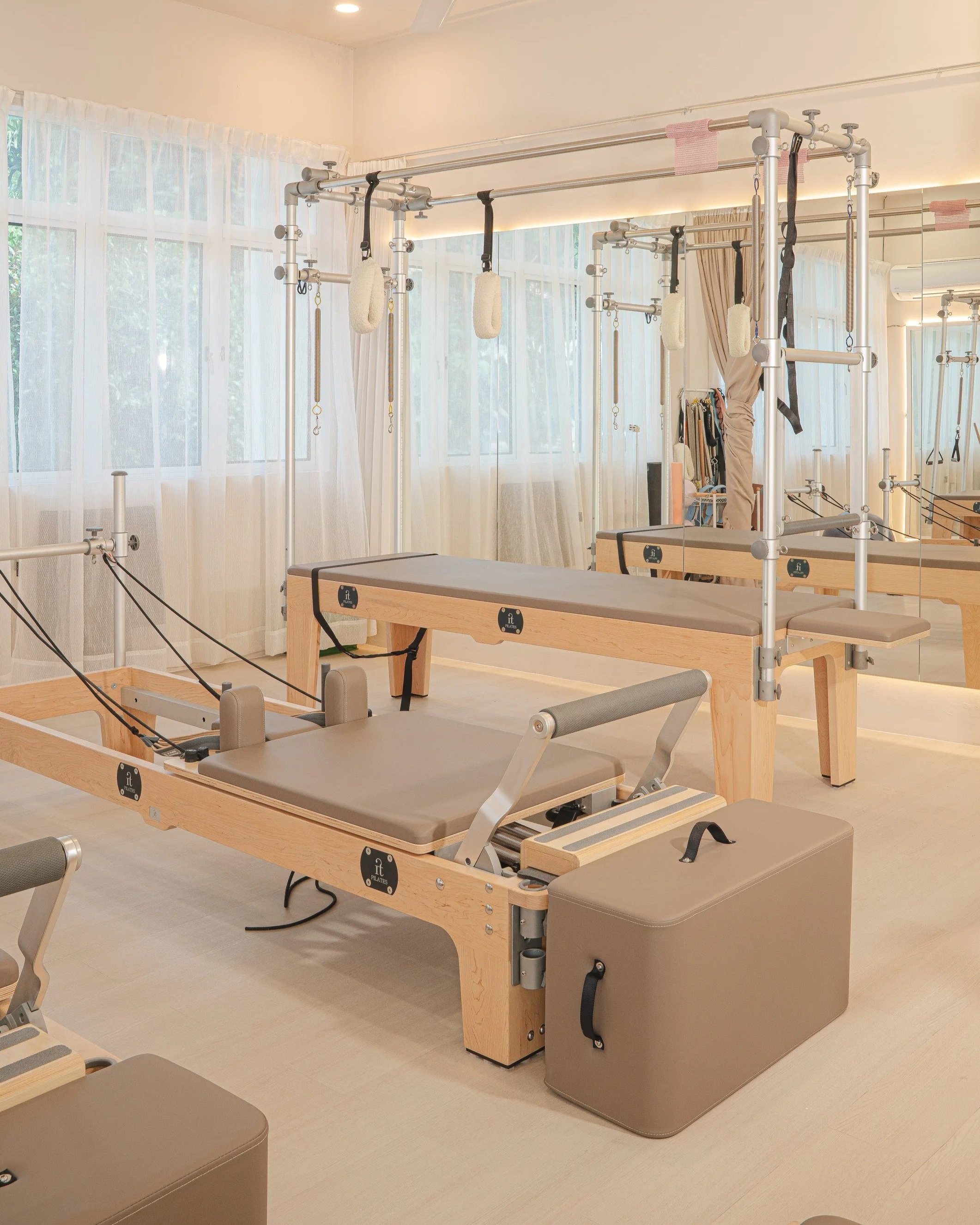 A Pilates studio with wooden equipment, padded benches, and suspension straps in front of large windows with sheer curtains.