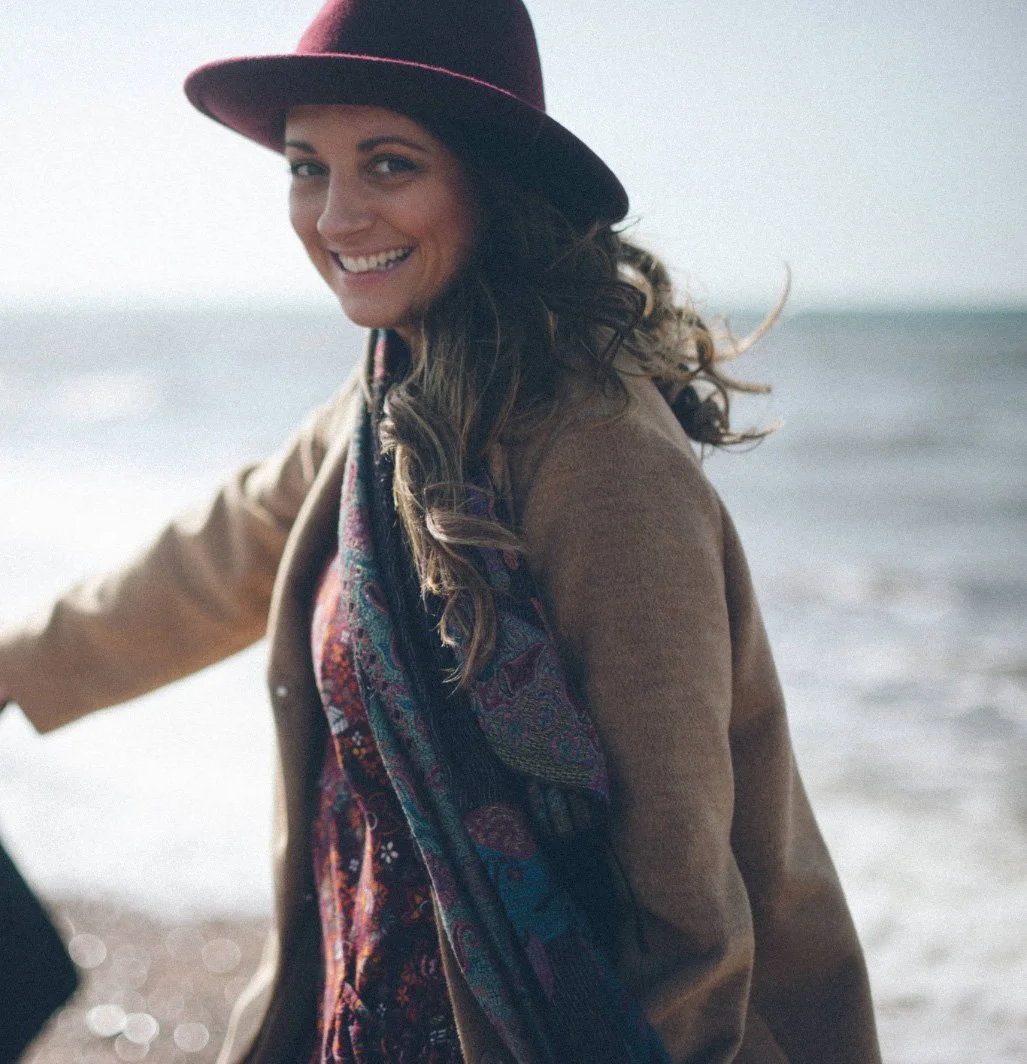 Amie Duke-Woolley, founder of Wild Mind Studios, on a windswept beach wearing a burgundy hat and patterned scarf