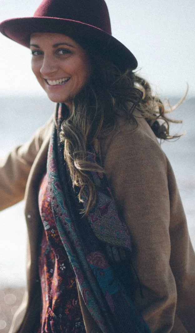 Amie, founder of Wild Mind Studios, smiling on the beach in a burgundy hat and paisley scarf