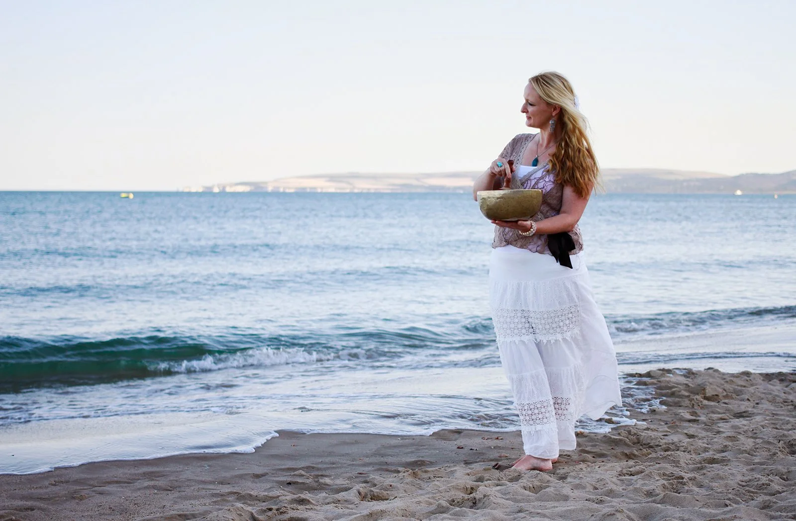 Woman in white lace dress standing by the sea holding a singing bowl — personal branding photography for Emma Penman by Wild Mind Studios