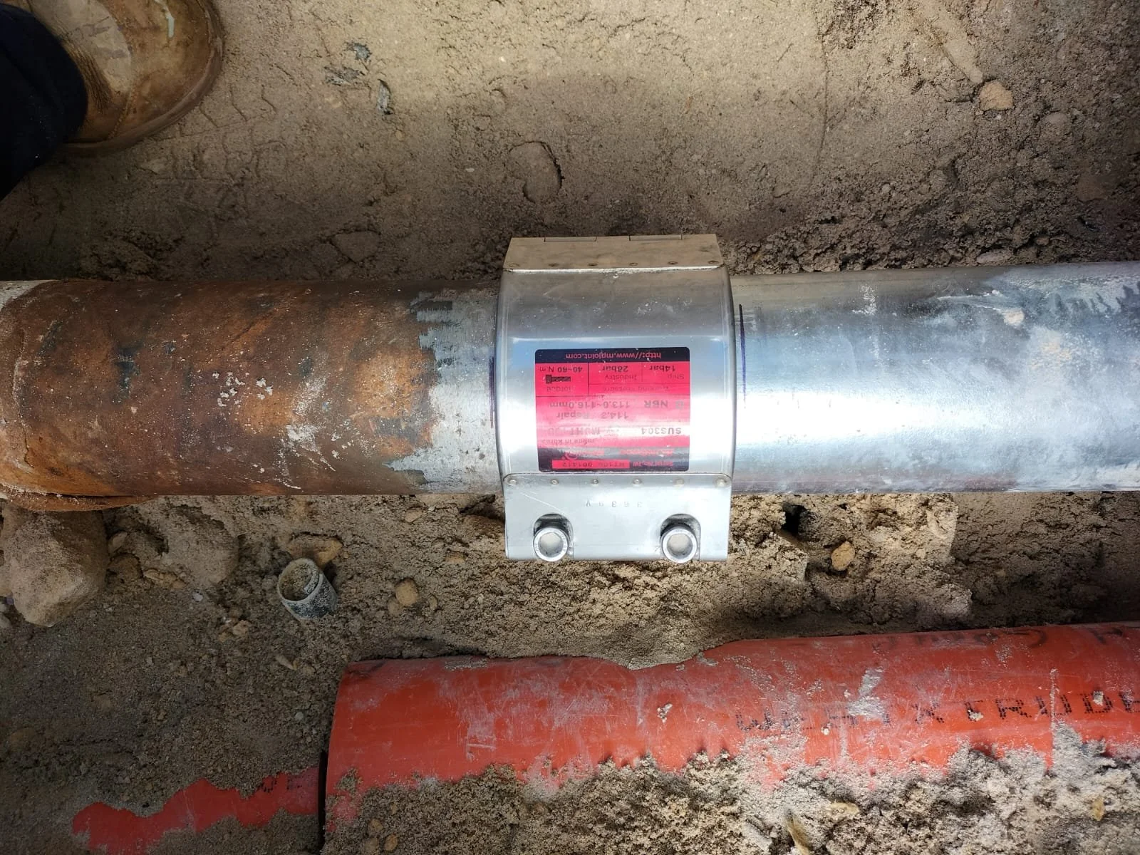 A metal pipe with a label clamp attached, surrounded by dirt and orange pipes at a construction site.
