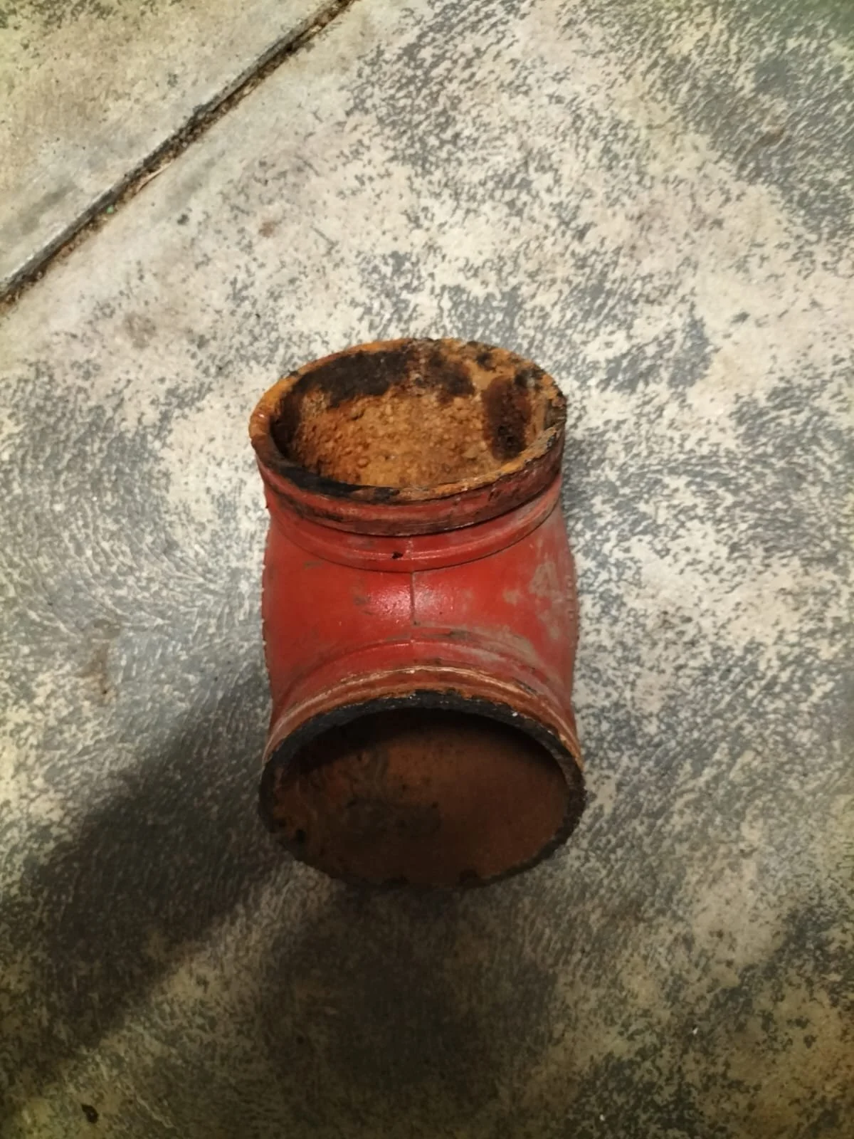 A rusted red pipe fitting, likely a T-joint, with burned interior showing soot and corrosion, sitting on a textured gray concrete floor.