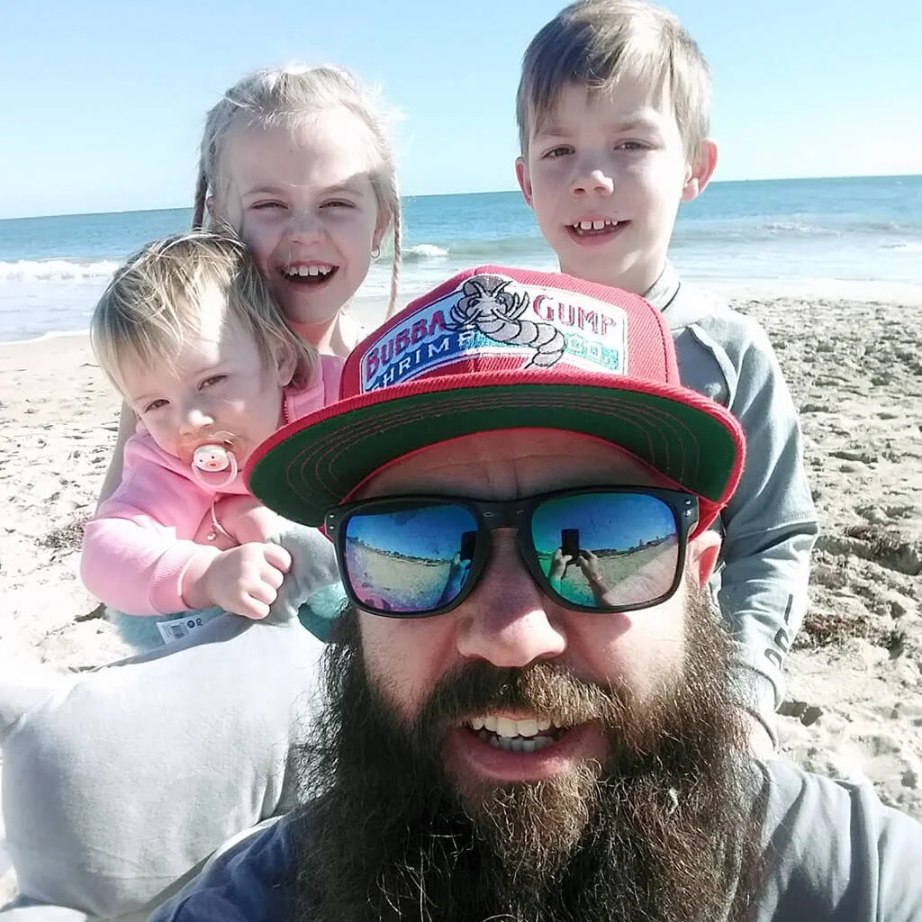 A bearded man wearing sunglasses and a red cap with a cartoon character taking a selfie on a sandy beach with three children, the ocean in the background.