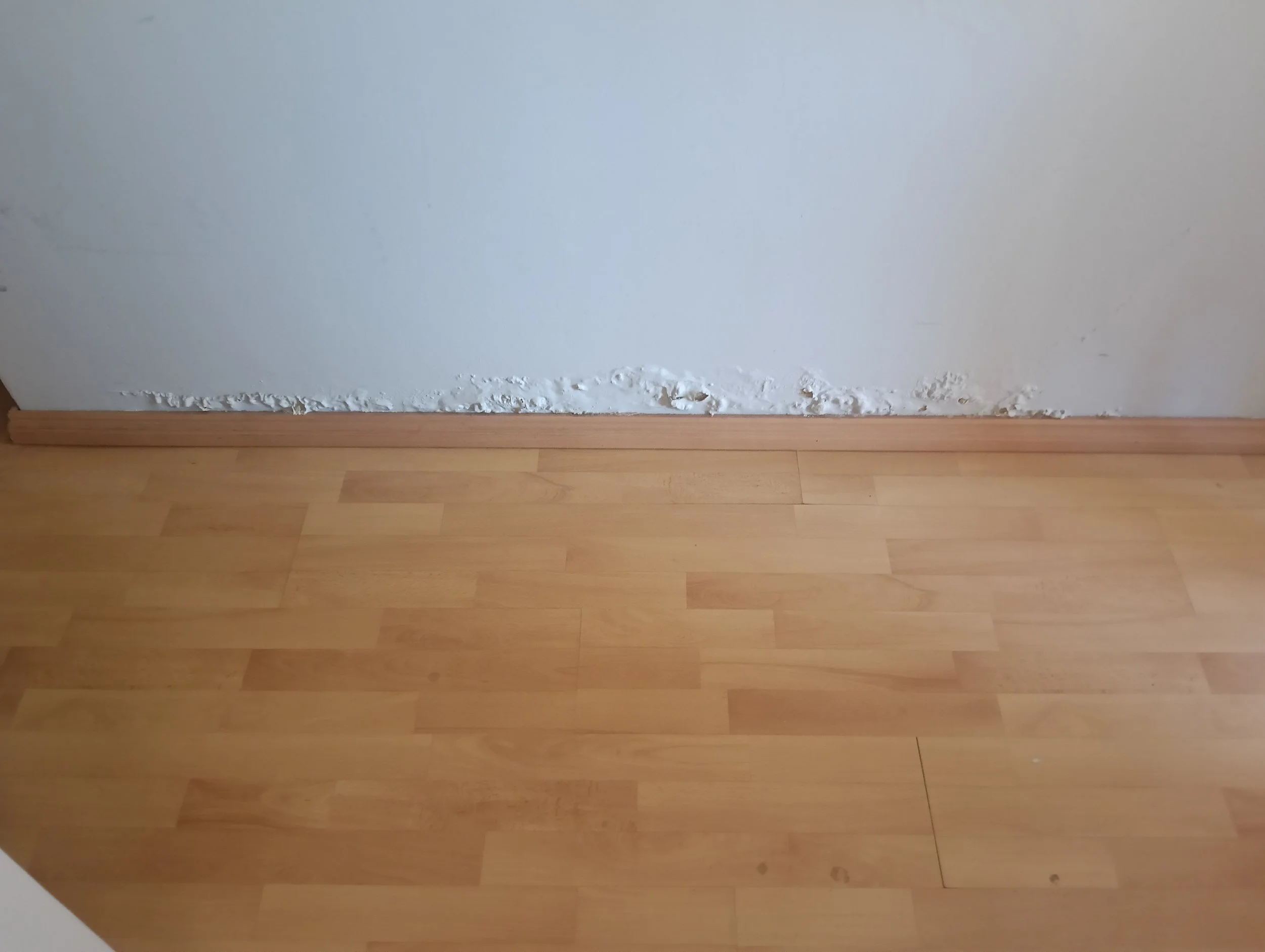 Baseboard along the bottom of a wall with damaged and peeling paint above a brown wooden floor.