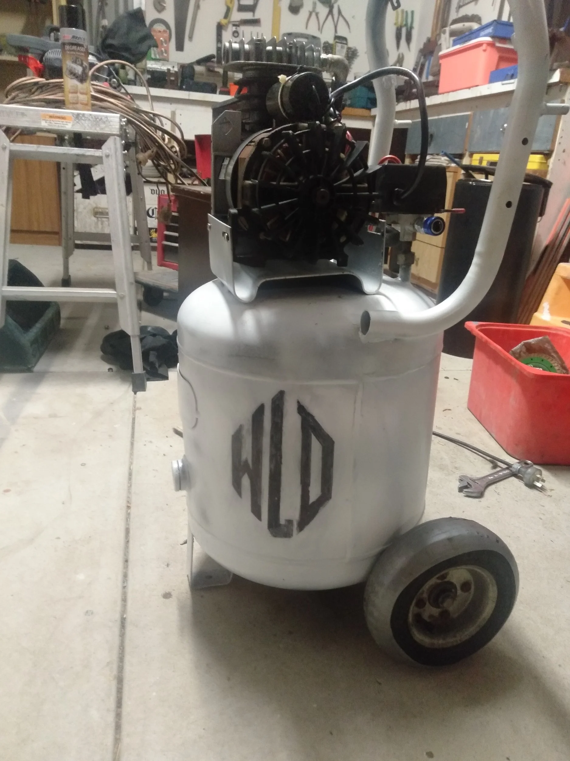 A homemade or customized air compressor cart with a white tank, black logo, and a compressor mounted on top, set in a workshop with tools and equipment in the background.