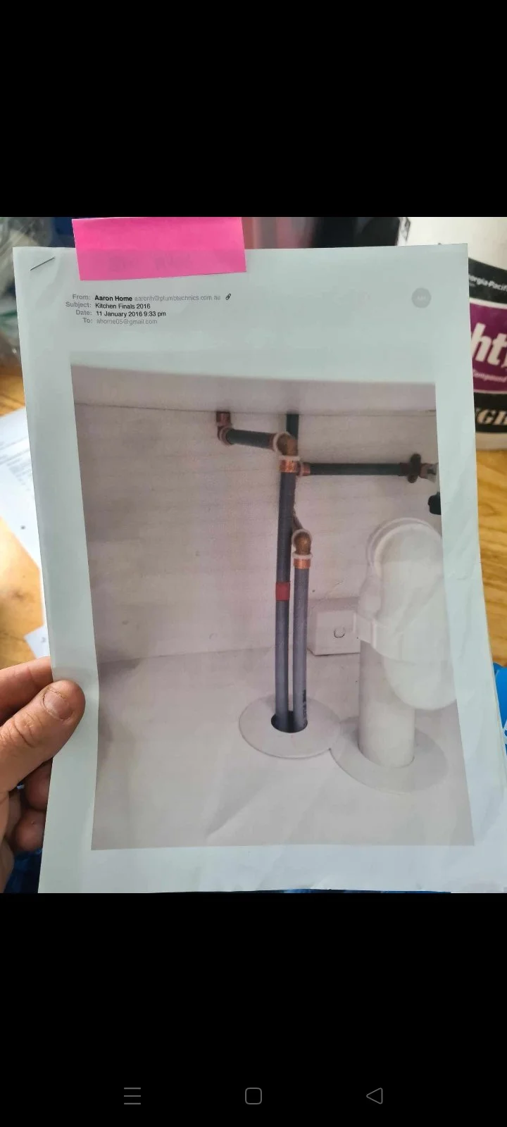 Photograph of a printed photo showing plumbing pipes and fittings installed through a wall, with a person's hand holding it up. The pipes are grey with copper fittings, situated near a white wall.
