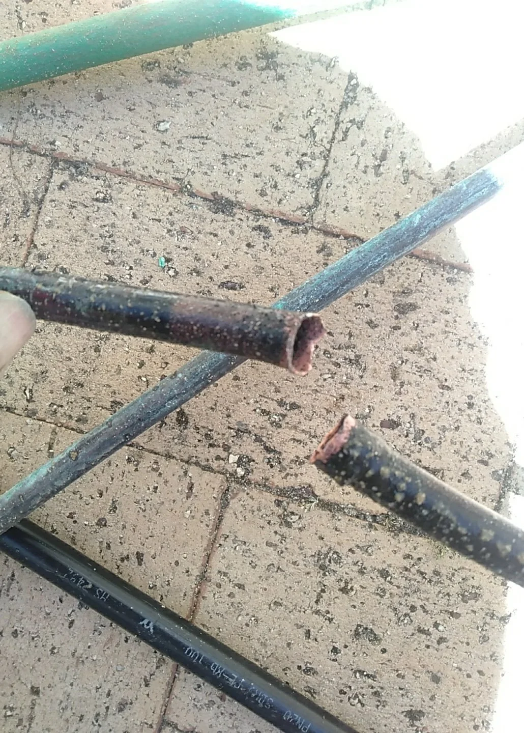Close-up view of rusty, hollow metal pipes on a brick patio, some broken at the ends, with dirty dust and debris on the ground.