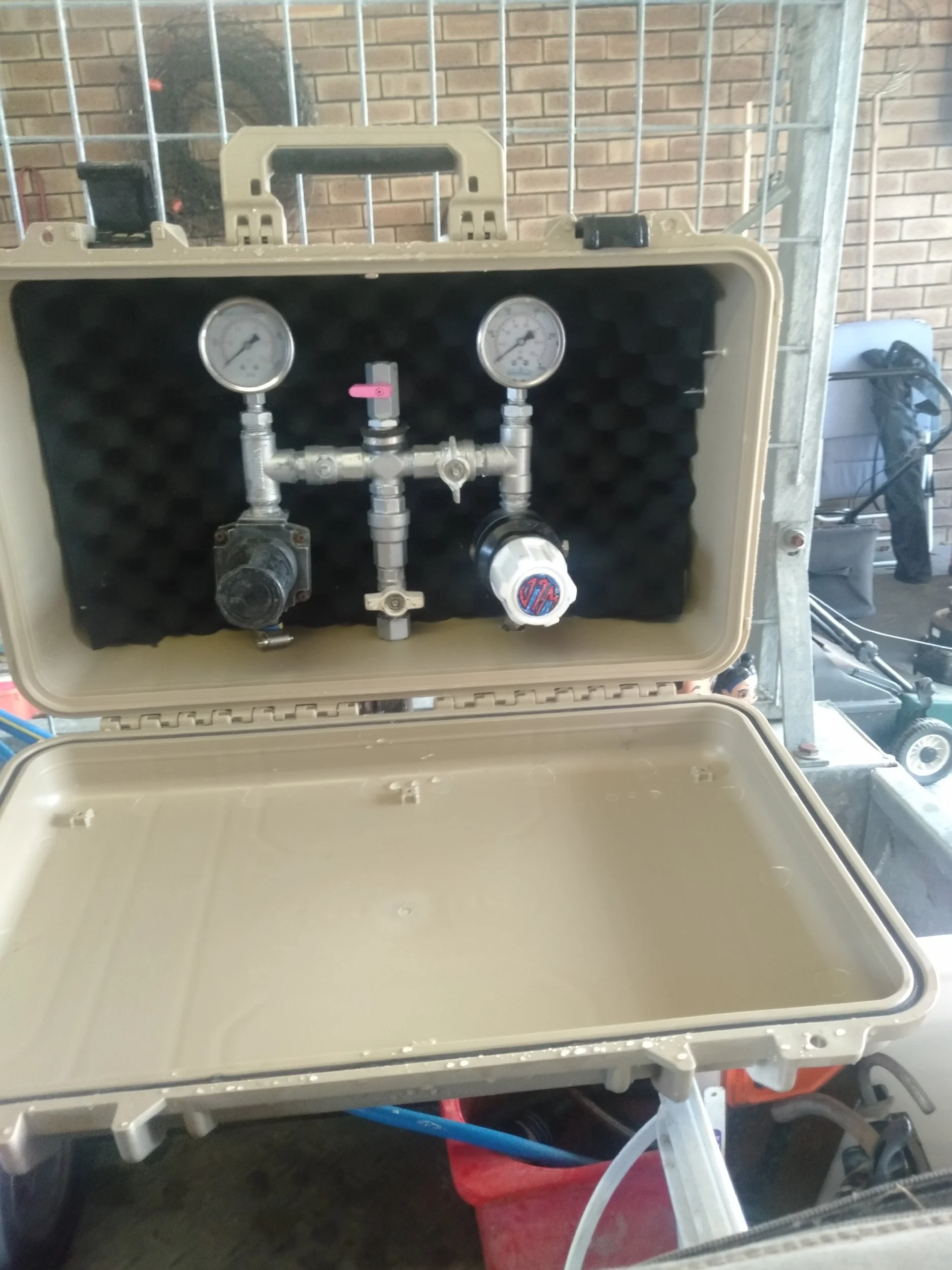 Closed beige plastic case with black foam lining in the lid, containing two pressure gauges, a valve, and plumbing fittings.