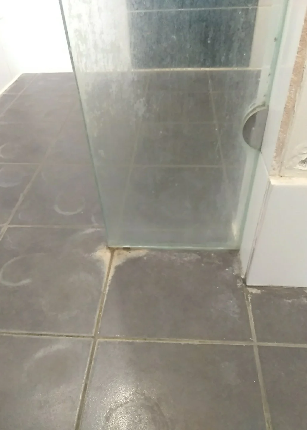 Close-up of a glass shower enclosure corner with black tiled floor and some dirt or soap scum around the base.