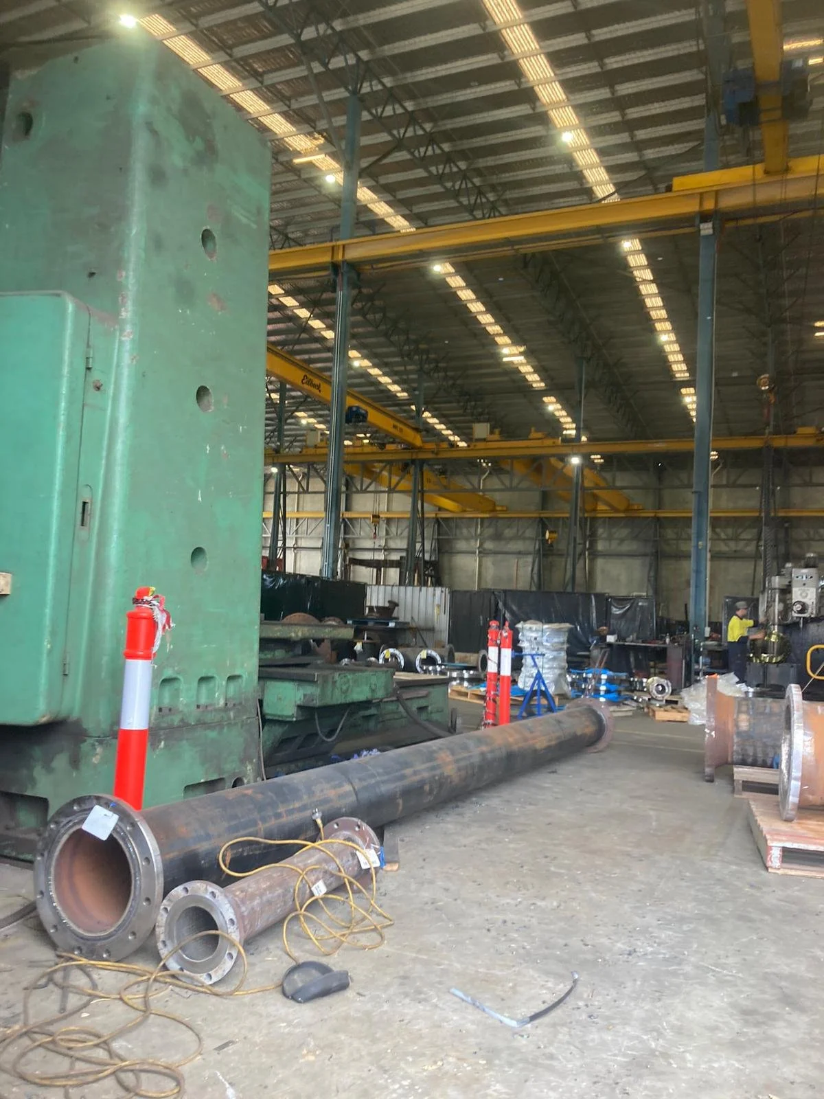 Inside an industrial warehouse with large metal pipes, machinery, and a worker, featuring yellow overhead cranes.