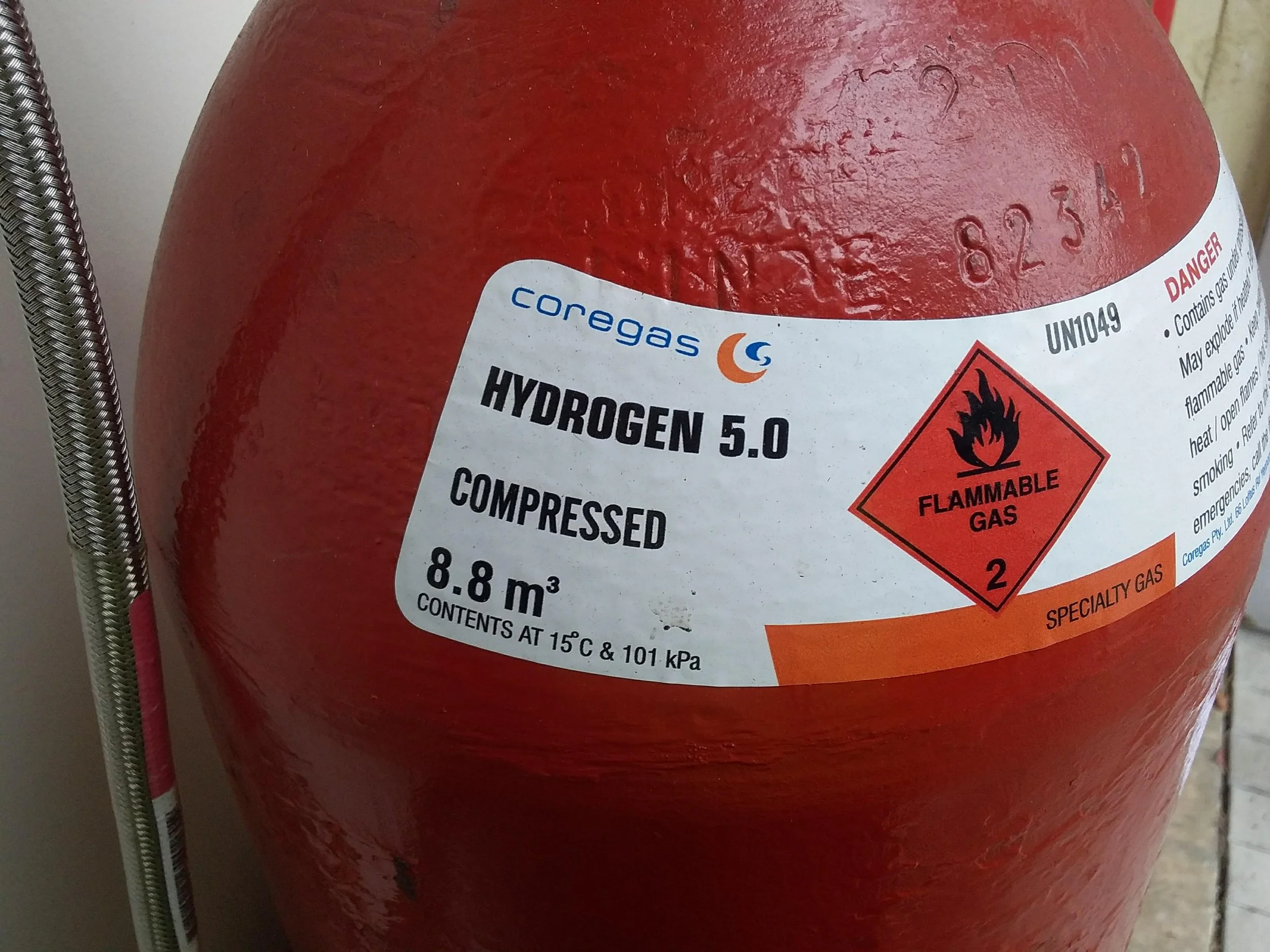 Red compressed hydrogen gas cylinder with a label indicating it contains 8.8 cubic meters of hydrogen at 15°C and 101 kPa, with flammable gas warning symbols.