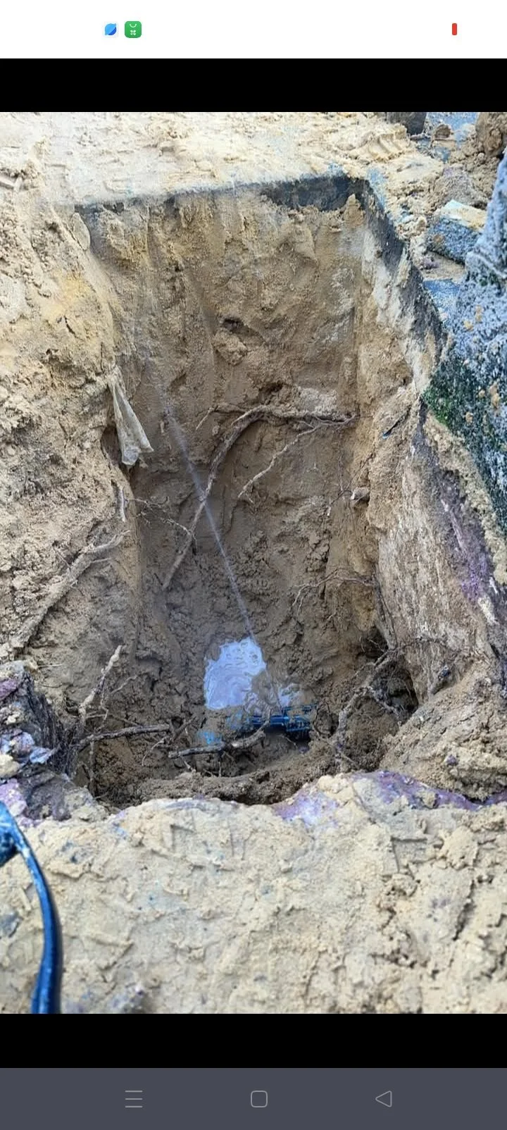 A deep excavation in the ground showing exposed roots and soil, with a small pool of water at the bottom. There's a device attached to wires at the bottom and some cables nearby.