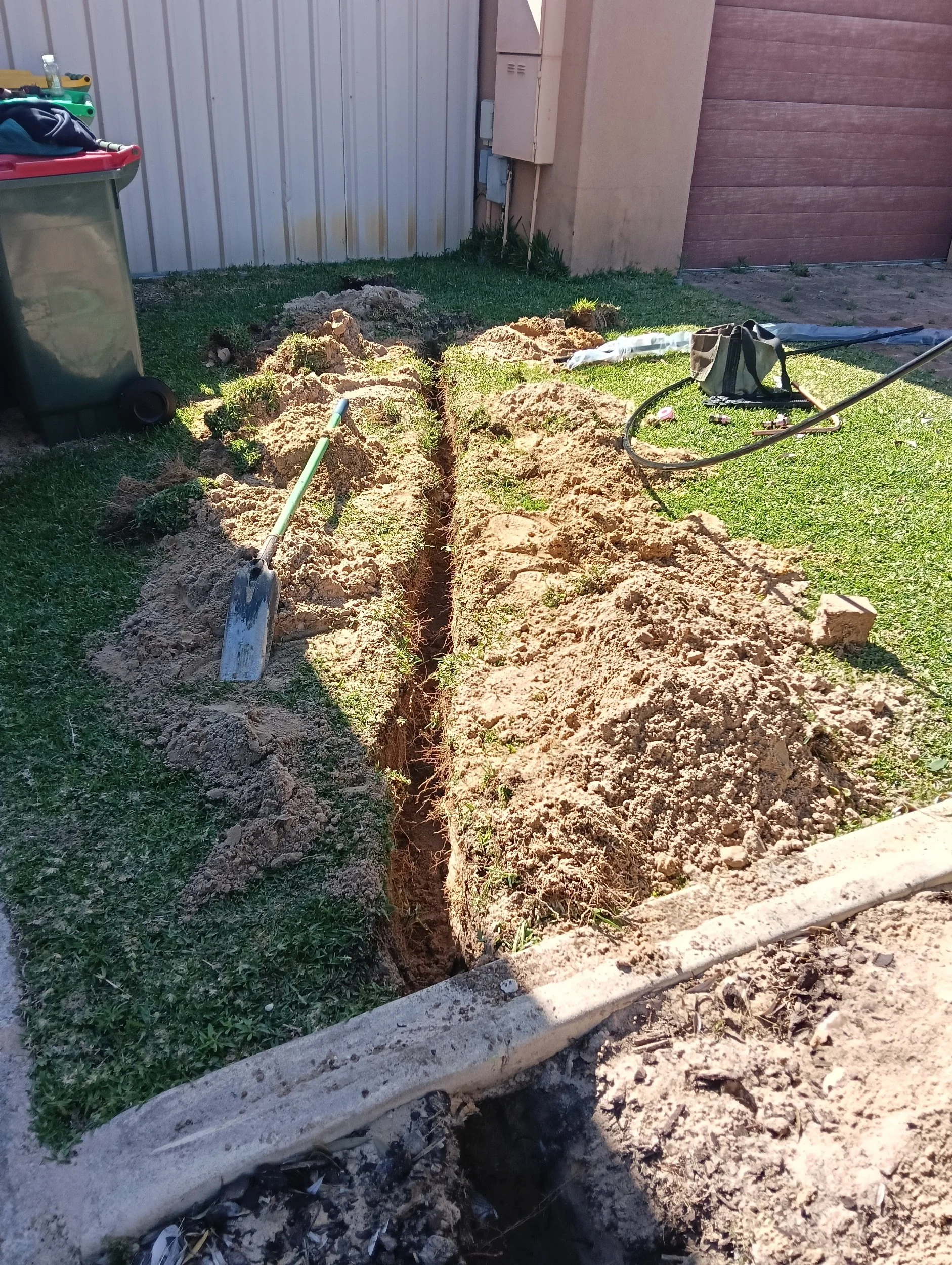 A trench is being dug in a yard with tools and equipment around, including a shovel and a garden hose. The trench runs along a grassy area next to a shed or garage.