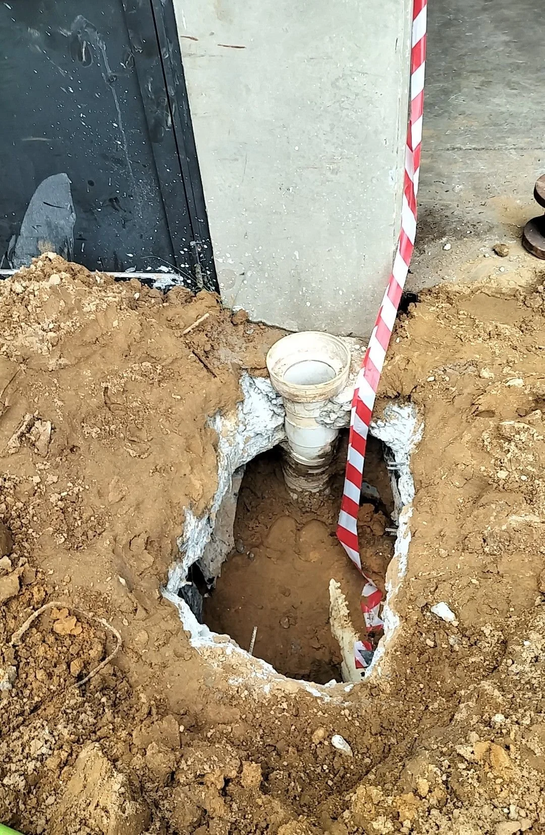 An open excavation with a visible pipe sticking out of the ground near a building foundation, surrounded by dirt and construction tools.