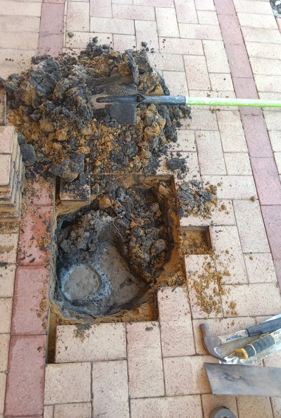 Digging up a square section of sidewalk with a shovel, revealing the hole and water or mud inside, with paving bricks and tools nearby.