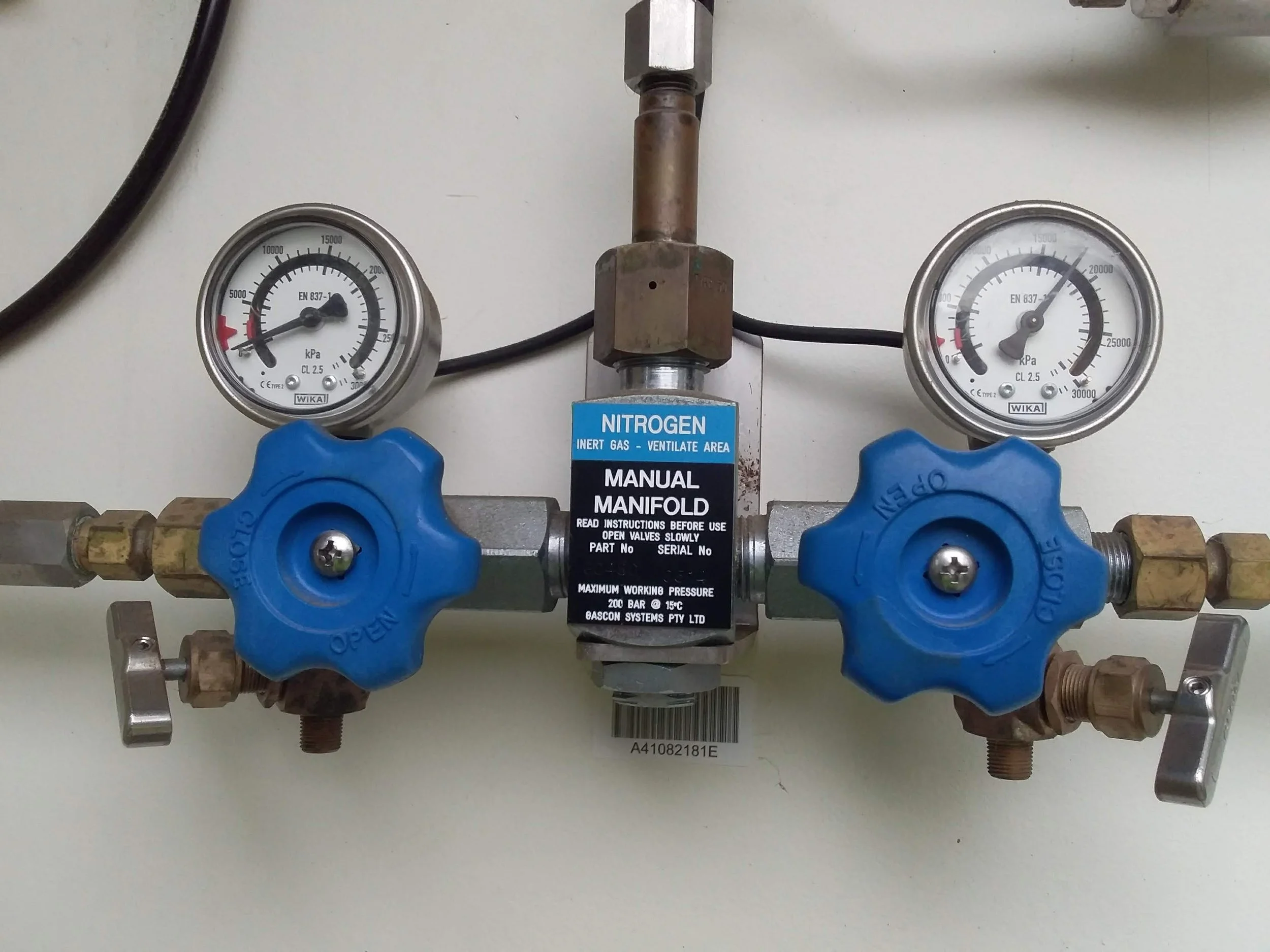 A nitrogen gas manifold with two pressure gauges, blue valve handles, and a manual valve in the center, mounted on a white surface.