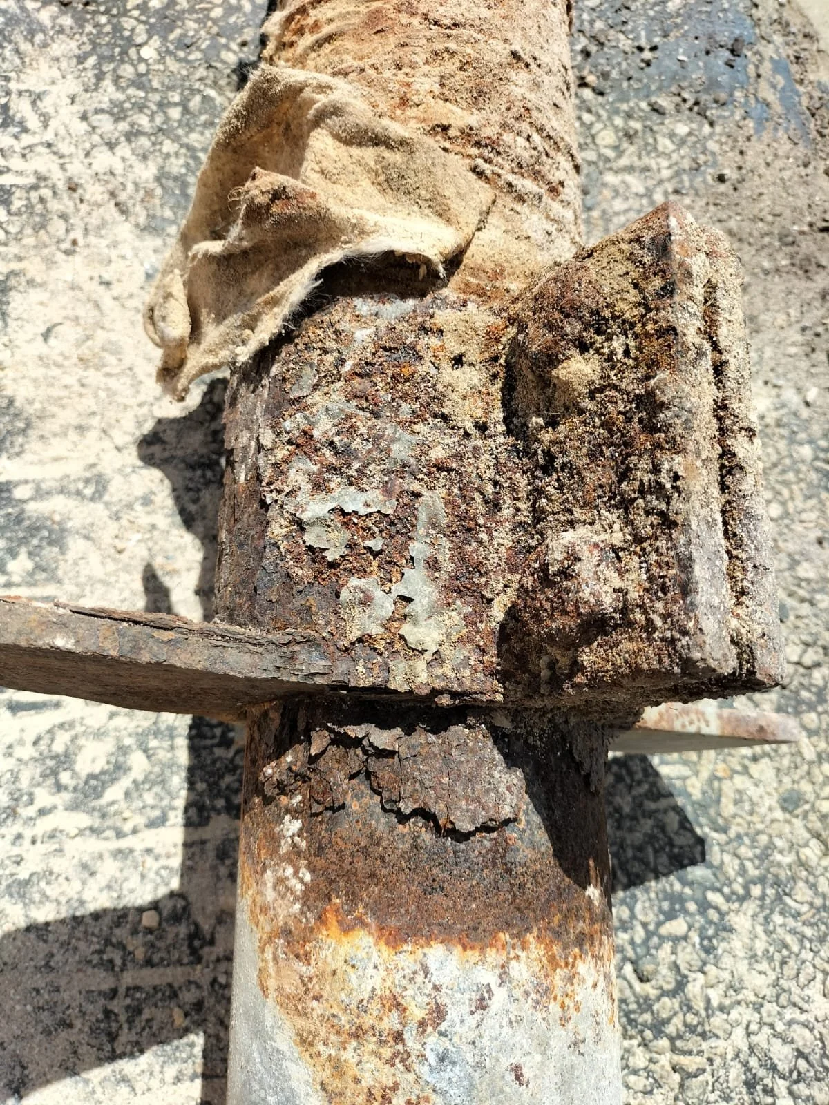 Close-up of a rusty, weathered metal object with peeling paint, attached to a rusted pipe or pole. It appears to be a piece of industrial equipment or debris.