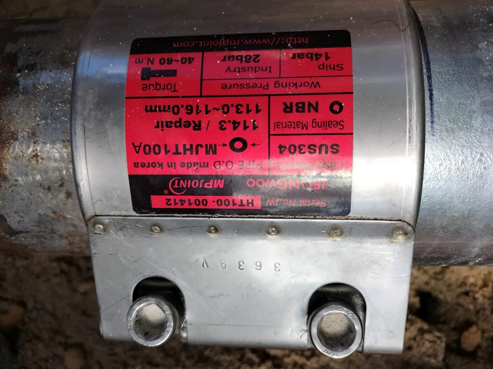Close-up of a metallic hydraulic component with a red and black label containing technical specifications, and two bolts at the bottom, on a dirt surface.