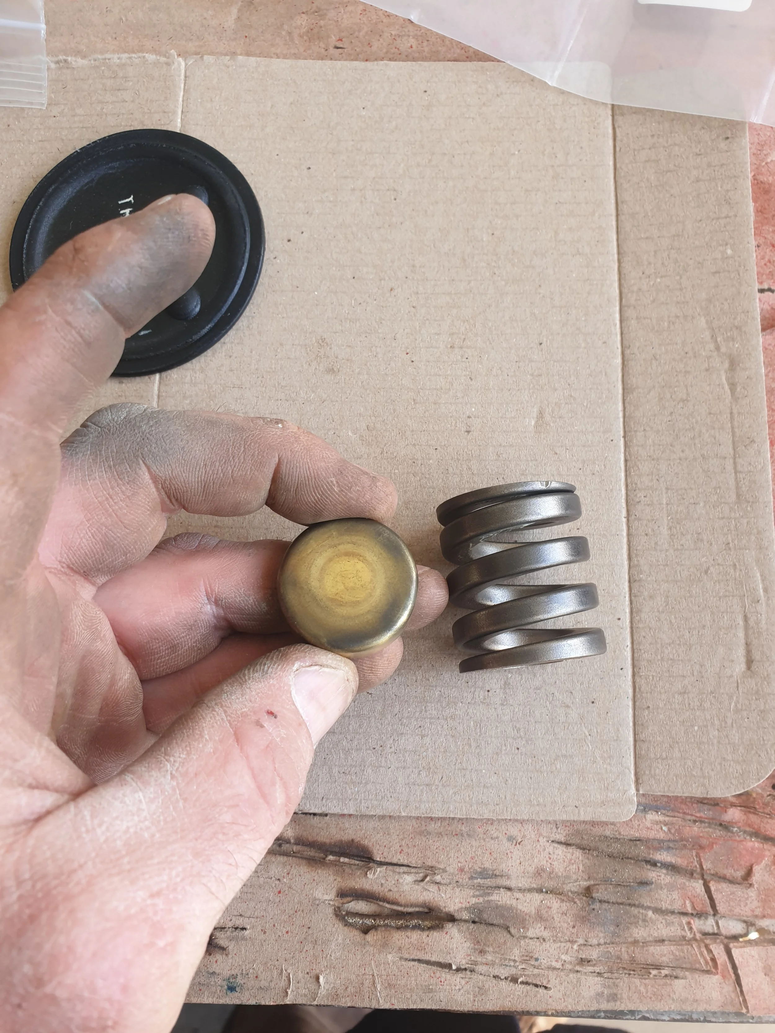 A hand holding a worn metal object, with a stack of similar metal objects and a black rubber piece on a cardboard surface.