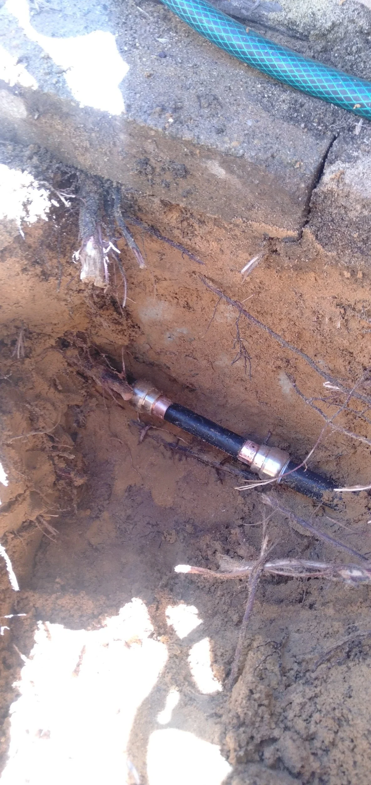 Close-up of a buried underground pipe with a clamp, exposed in a dirt trench, alongside a garden hose.